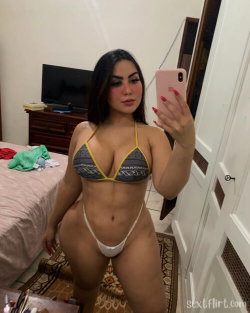 Curvy woman with dark hair takes a mirror selfie in a patterned bikini top and white thong, standing in a bedroom.