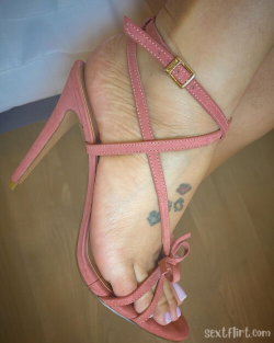 Close-up of a foot in pink strappy high heels with a small floral tattoo on the arch, standing on a wooden floor.