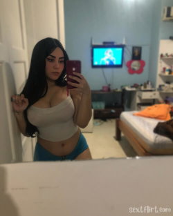 Brunette woman in a white crop top and blue shorts takes a mirror selfie in a bedroom with a TV in the background.