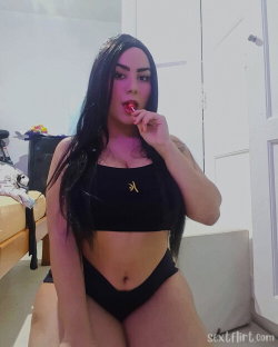 A woman with long dark hair poses in a black crop top and matching bottoms, holding a red lollipop in a bedroom setting.