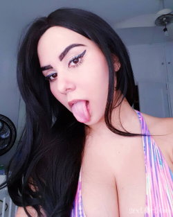 Brunette woman with long dark hair and bold eyeliner sticks out her tongue in a playful selfie, wearing a colorful