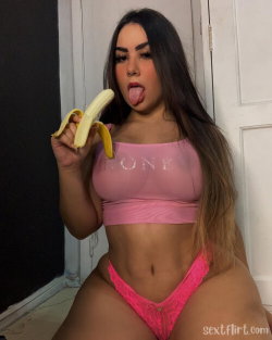 Curvy woman with long dark hair wearing a pink crop top and bright pink lace thong, holding a banana while sticking out