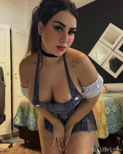 A woman with dark hair in a ponytail wears a plaid halter top and matching skirt, posing indoors with hands on thighs.