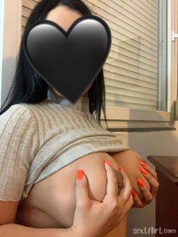 Brunette woman with curvy build wearing a ribbed top, holding her bare breast with bright orange nails indoors near a