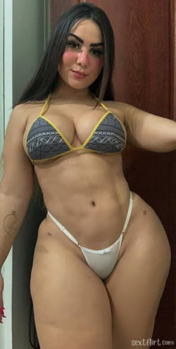 Brunette woman with curvy figure wearing patterned yellow-trimmed bikini top and white thong, posing indoors near a