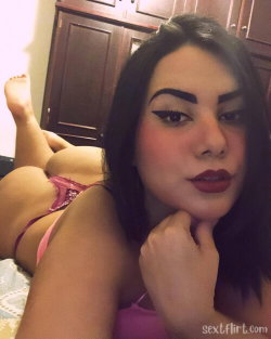 A curvy woman with dark hair and bold makeup lies on a bed in pink lingerie, posing for a selfie with one leg raised.