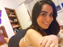 Brunette woman with a curvy figure wearing black shorts and a white bra, lying on a bed in a bedroom, smiling at the