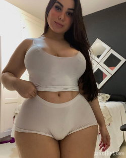 Brunette woman with curvy figure wearing white crop top and matching high-waisted underwear, posing in a bedroom.
