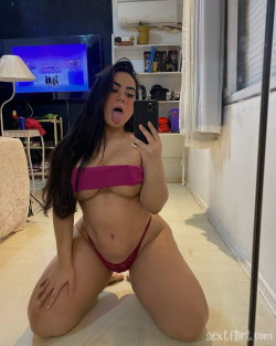 Brunette woman with curvy figure in pink bandeau top and red thong, kneeling on floor taking mirror selfie in living