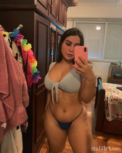 Curvy woman in a gray top and blue thong takes a mirror selfie in a bedroom with wooden furniture and colorful decor.