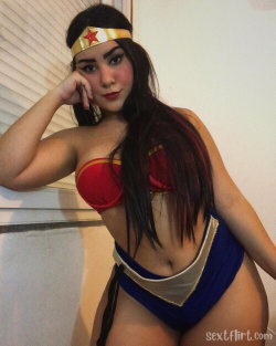 A woman with long dark hair in a Wonder Woman costume poses indoors, wearing a red bra top and blue skirt with gold