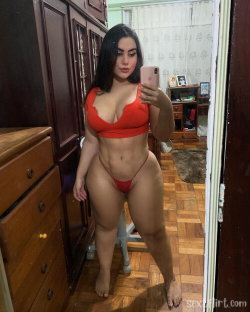 A woman with dark hair in a red bikini takes a mirror selfie in a bedroom with wooden furniture and parquet flooring.