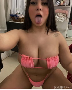 A curvy woman with dark hair takes a selfie in a pink bikini top and thong, sticking her tongue out in a playful pose