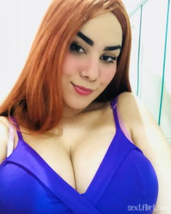 Redheaded woman with defined eyebrows and a nose ring wears a deep V-neck blue top, posing closely in a selfie-style