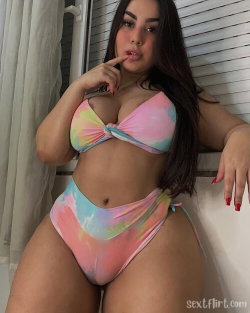 Curvy woman with long dark hair wearing a colorful tie-dye bikini, posing indoors near a window with one hand on her