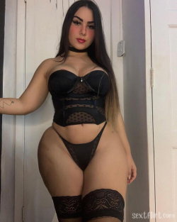Curvy woman with long dark hair wearing black lace lingerie and garter straps, posing indoors near a white door.