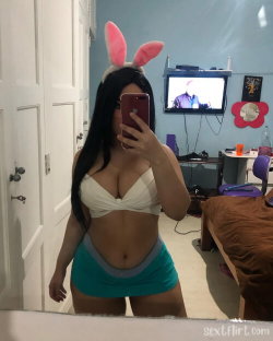 A woman with long dark hair wearing pink bunny ears, a white crop top, and teal shorts takes a mirror selfie in a