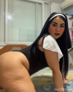 A woman with long dark hair in a black and white nun-style outfit bends forward, showing her backside in a bedroom