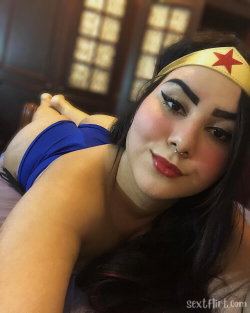 Brunette woman in a Wonder Woman costume with blue top and gold headband, lying on her side indoors with dramatic
