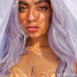 A woman with long lavender hair and a sheer veil, wearing a delicate necklace, with dappled sunlight on her face and