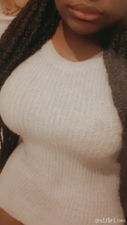 Close-up of a woman with long dark curly hair wearing a tight white knit top, showing her upper body and chest area.