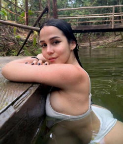 Brunette woman in white bikini top leaning on wooden dock in calm water, surrounded by lush greenery.