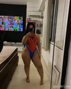 Curvy woman in a Spider-Man bodysuit takes a mirror selfie in a bedroom with a bed and TV visible.