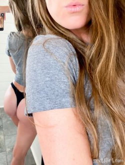 A woman with long brown hair in a gray t-shirt takes a selfie, showing her back and side profile with another person