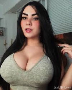 A woman with long dark hair and a curvy build wears a tight olive-green top, posing indoors with one hand in her hair.
