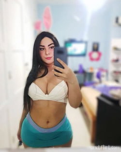 A woman with long dark hair takes a mirror selfie in a white top and teal skirt, wearing pink bunny ears in a bedroom