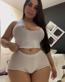 Curvy woman with long dark hair wearing white crop top and matching high-waisted underwear, posing in a bedroom.