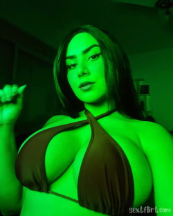 A woman with dark hair and a nose ring wears a brown halter top under green lighting, posing in a dimly lit room.