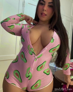 Curvy woman with long dark hair wearing a pink avocado-print onesie, posing indoors with one hand near her neck.