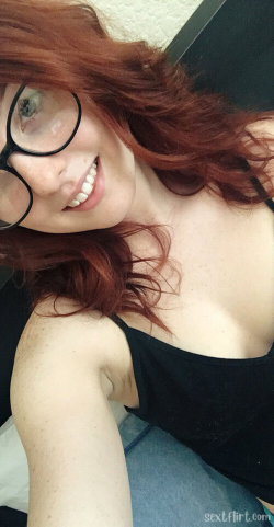 A woman with red hair and glasses smiles in a close-up selfie, wearing a black tank top and blue jeans indoors.