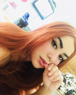 A woman with long red hair and a nose ring poses playfully in a selfie, touching her lips with her hand indoors.