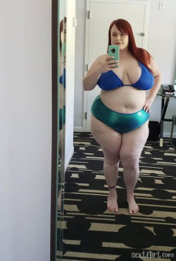 Redheaded woman in a blue bikini top and teal mermaid-patterned bottoms takes a mirror selfie in a hotel room.