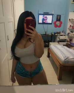A woman with long dark hair takes a mirror selfie in a bedroom, wearing a white crop top and patterned shorts.