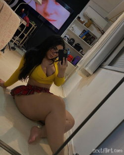 Brunette woman in yellow crop top and red plaid shorts poses on floor, taking mirror selfie in room with TV and shelves.
