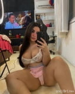 Curvy woman with long dark hair takes a mirror selfie in lace top and pink shorts, sitting on carpeted floor in a room