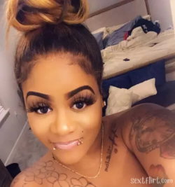 A woman with dark skin and styled hair in a bun takes a selfie, showing tattoos on her chest and arm, wearing a