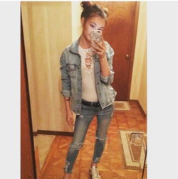 A young woman with dark hair in a bun takes a mirror selfie, wearing a denim jacket, lace-up top, ripped jeans, and