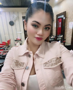 A woman with dark hair and makeup takes a selfie in a pink jacket over a white top, indoors in what appears to be a