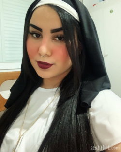 Woman with long dark hair wearing a nun-style hood and white top, posing indoors with makeup and a nose ring.