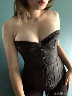 Woman with shoulder-length hair wearing a black lace corset, standing against a blue background.