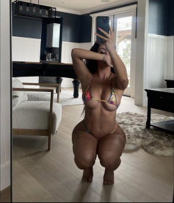Curvy woman in colorful bikini crouches for mirror selfie in a modern living room with navy walls and light wood floors.