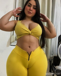 Curvy woman with long dark hair wearing a yellow crop top and matching high-waisted pants, posing indoors with hands in