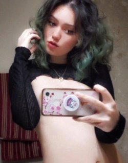 A young woman with dark hair and green highlights takes a mirror selfie, wearing a black long-sleeve top pulled down to
