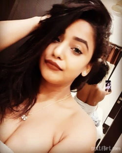 A woman with dark hair and a bare shoulder takes a mirror selfie, wearing a delicate necklace and white top, in a