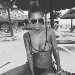 A woman with tattoos on her arms and shoulders wears a mesh bikini top and sunglasses, sitting under a thatched