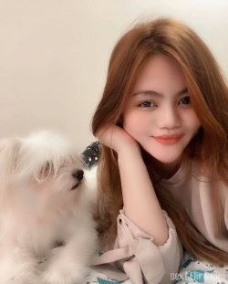 A young woman with long brown hair smiles softly while resting her head on her hand beside a small white dog, wearing a
